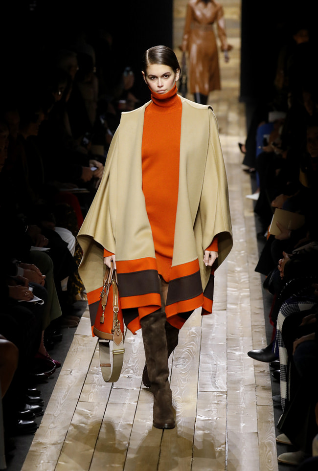 Kaia Gerber rules the runway in an oversized layered shawl and a dark orange sweater. The orange sweater is a perfect contrast to the neutral colors of the shawl.
